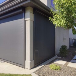 House corner with black roller shades, green bushes nearby.